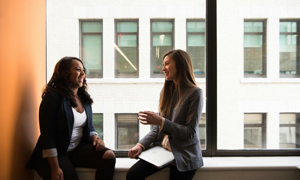two women in business attire sitting and talking in an office