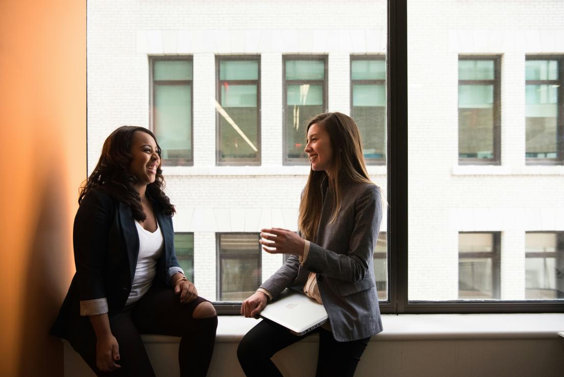 two women in business attire sitting and talking in an office