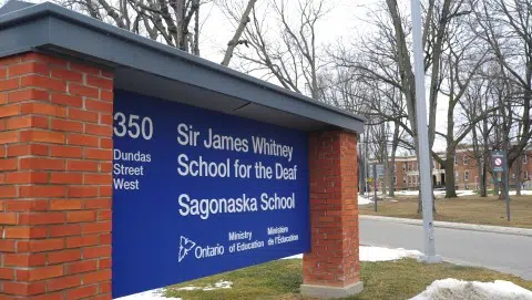A blue sign at the entrance of Sir James Whitney School for the Deaf and Sagonaska School in Ontario, with buildings and bare trees visible in the background.