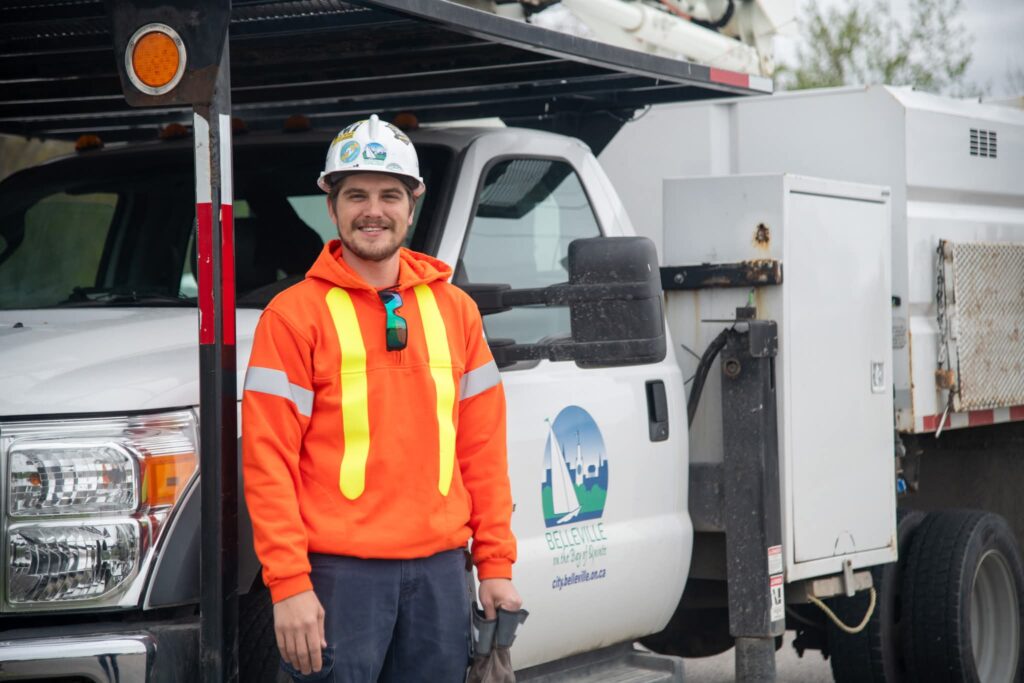 A construction worker, working in the trades, wears a neon orange safety jacket and helmet as he stands next to a white utility truck with city branding on the door.