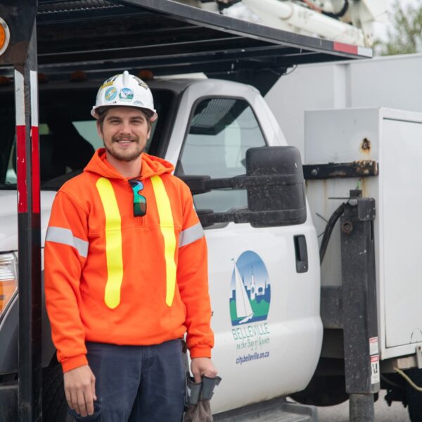 A construction worker, working in the trades, wears a neon orange safety jacket and helmet as he stands next to a white utility truck with city branding on the door.