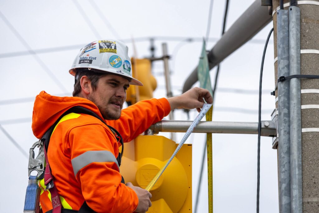 A construction worker, working in the trades, wears an orange safety jacket and hard hat while measuring a traffic light pole with a tape measure.