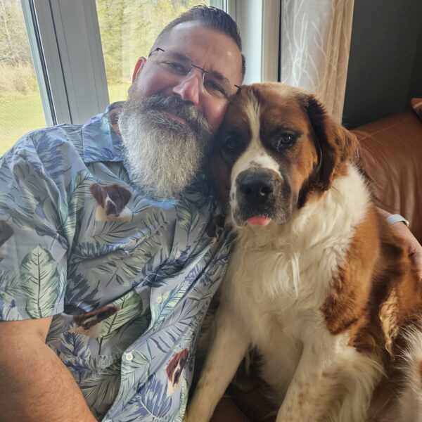 A man with a gray beard sits on a couch next to a large Saint Bernard dog, both facing the camera, while the cozy setting and soft lighting reflect the warm ambiance of Aesthetics by Lindsay Ann.
