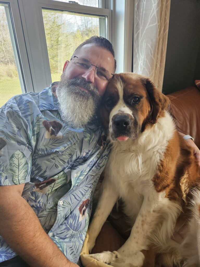 A man with a gray beard sits on a couch next to a large Saint Bernard dog, both facing the camera, while the cozy setting and soft lighting reflect the warm ambiance of Aesthetics by Lindsay Ann.