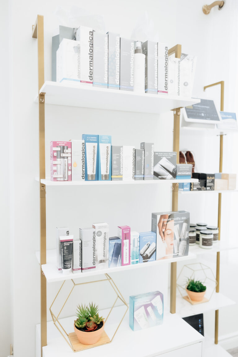 Three white shelves display various Dermalogica skincare products in boxes at Aesthetics by Lindsay Ann. Two small potted succulents sit on the bottom shelf, and light enters from the right.