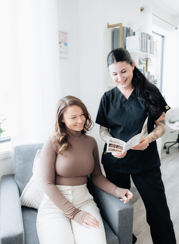 A woman sits in a chair while a smiling healthcare professional from Aesthetics by Lindsay Ann in scrubs stands beside her, holding and showing her a brochure.