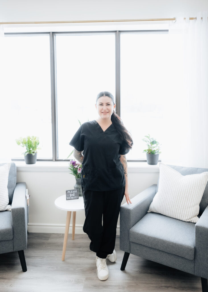 A woman in black scrubs, working in the trades, stands smiling between two gray chairs in a bright room with large windows, potted plants, and a small round table.