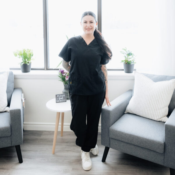 A woman in black scrubs, working in the trades, stands smiling between two gray chairs in a bright room with large windows, potted plants, and a small round table.