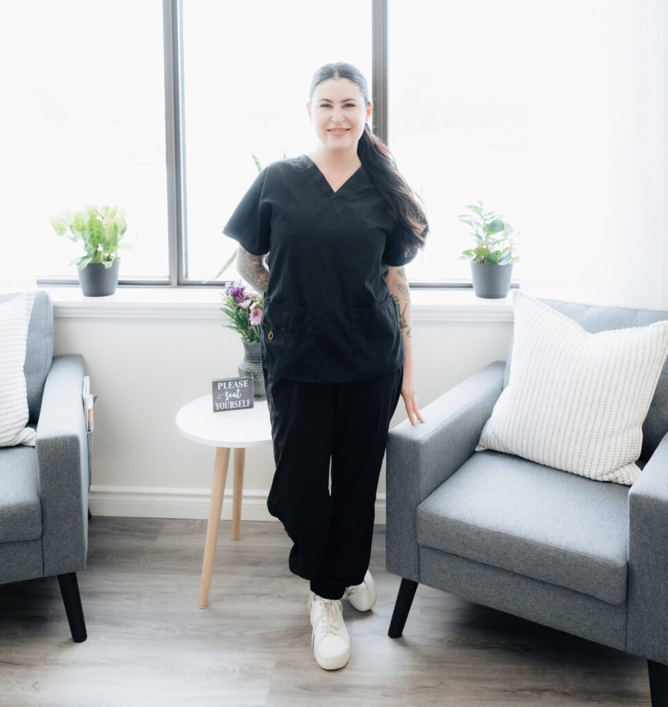 A woman in black scrubs, working in the trades, stands smiling between two gray chairs in a bright room with large windows, potted plants, and a small round table.