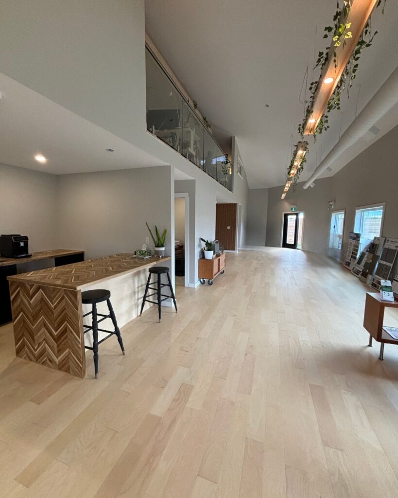 Spacious modern interior by Simple Life Homes with light wood flooring, a wooden reception desk and two stools, glass railing on the upper level, and plants hanging from the ceiling.