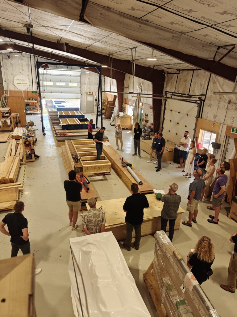 A group of people gather around a speaker in a warehouse with construction materials and wooden frames laid out on the floor, discussing designs for Simple Life Homes.
