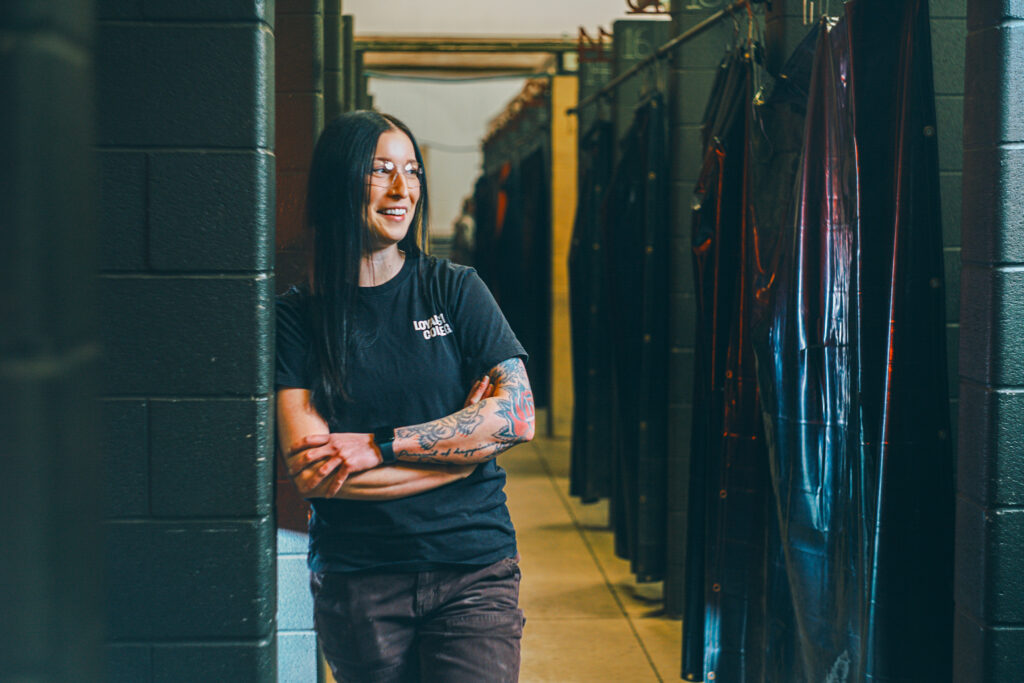 A person working in the trades with long dark hair, glasses, and tattoos leans against a wall in an industrial hallway lined with dark hanging items, wearing a black t-shirt and pants.