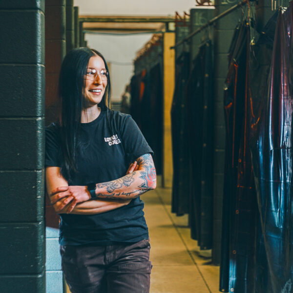 A person working in the trades with long dark hair, glasses, and tattoos leans against a wall in an industrial hallway lined with dark hanging items, wearing a black t-shirt and pants.