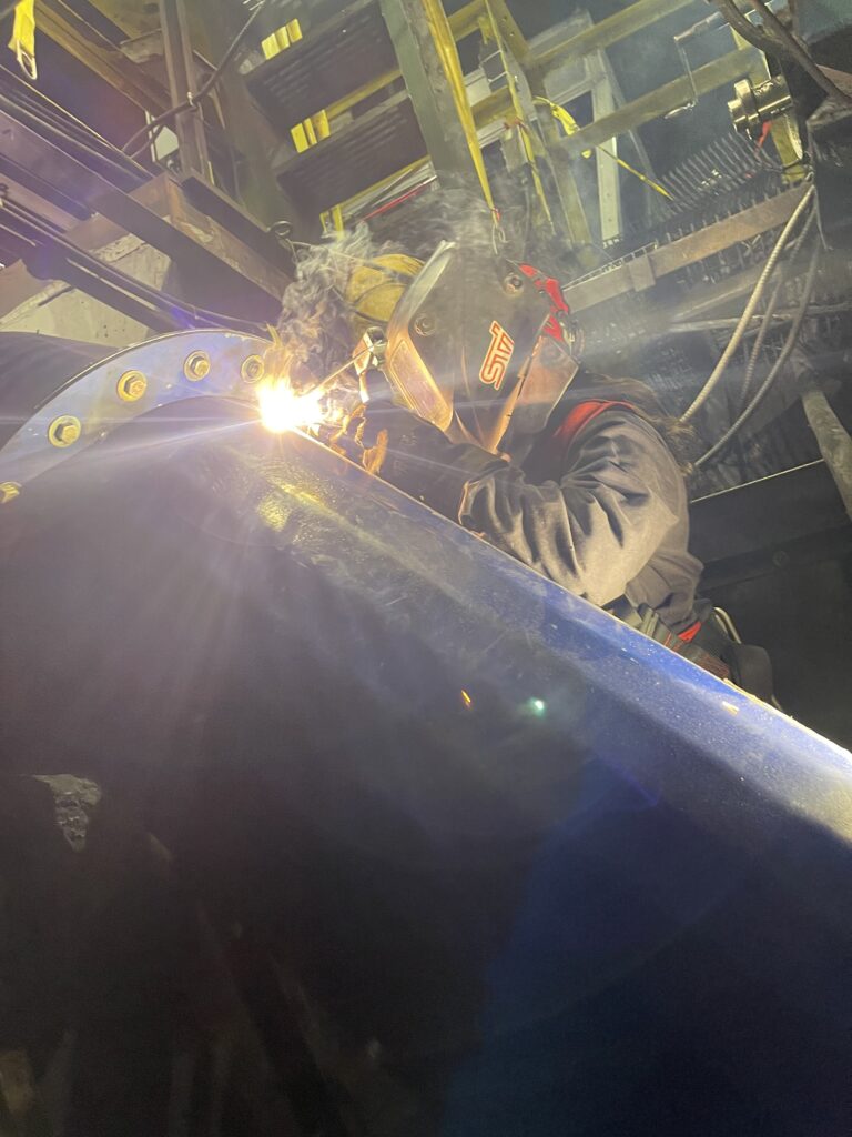 A person working in the trades, wearing protective gear and a welding mask, is welding metal beams in an industrial setting, with sparks and light visible.
