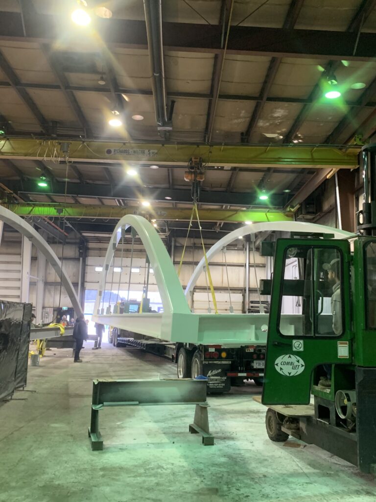 Large white metal arches are being lifted by a yellow overhead crane inside a warehouse, showcasing the skills of those working in the trades; a green forklift and flatbed truck stand nearby.