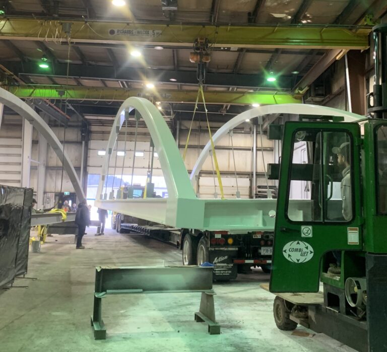 Large white metal arches are being lifted by a yellow overhead crane inside a warehouse, showcasing the skills of those working in the trades; a green forklift and flatbed truck stand nearby.