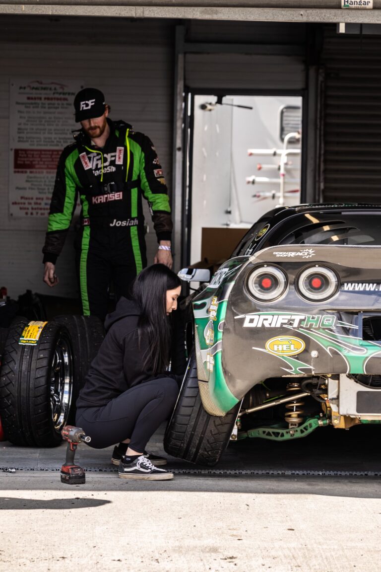 A woman kneels to inspect or adjust a rear tire on a drift car—her focus and skill echo those working in the trades—while a man in a racing suit stands nearby in the garage area.