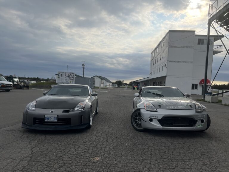 Two Nissan 350Z cars, perfect for those working in the trades, are parked side by side on an asphalt lot near a white building under a partly cloudy sky.