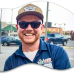 A man wearing sunglasses and a cap smiles outdoors while dressed in a blue collared shirt with badges on it; cars and buildings are visible in the background, showcasing the vibrant spirit of Bay of Quinte entrepreneurs.