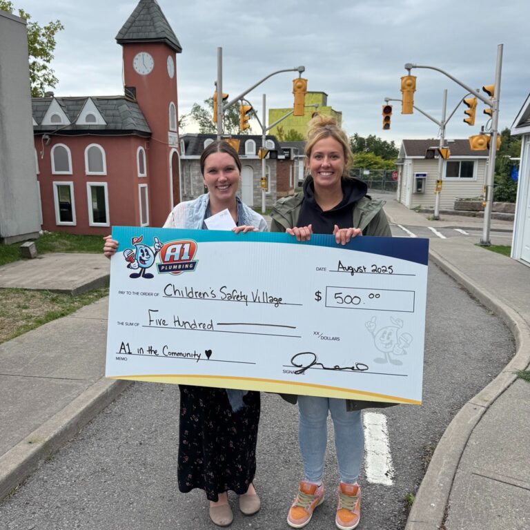 Two women stand outside holding a $500 check made out to Children's Safety Village from A1 in the Community, dated August 2023—showcasing the generosity of Bay of Quinte entrepreneurs.