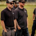 Two men, possibly Bay of Quinte entrepreneurs, wear sunglasses, black shirts, and caps as they stand outdoors on grass with hands in pockets; other people are partially visible in the background.