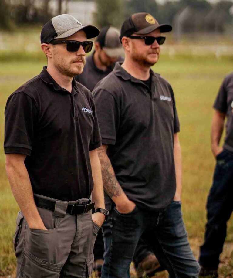 Two men, possibly Bay of Quinte entrepreneurs, wear sunglasses, black shirts, and caps as they stand outdoors on grass with hands in pockets; other people are partially visible in the background.