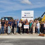 A large group, including Bay of Quinte entrepreneurs, poses in front of construction equipment and a sign at a building site. Some wear hard hats and safety vests, while two people in the center hold a “Thank You” sign under sunny blue skies.