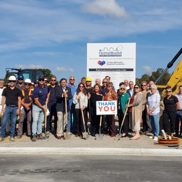 A large group, including Bay of Quinte entrepreneurs, poses in front of construction equipment and a sign at a building site. Some wear hard hats and safety vests, while two people in the center hold a “Thank You” sign under sunny blue skies.