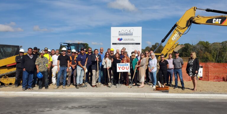 A large group, including Bay of Quinte entrepreneurs, poses in front of construction equipment and a sign at a building site. Some wear hard hats and safety vests, while two people in the center hold a “Thank You” sign under sunny blue skies.