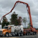 An orange and white concrete pump truck with an extended boom arm is parked at a construction site, surrounded by trees and a cloudy sky. A large signboard on the left highlights Bay of Quinte entrepreneurs contributing to the project.