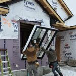 Two construction workers from Tobey Developments install a large window into the frame of a snow-covered house wrapped in Tyvek sheeting, with a green ladder propped nearby.