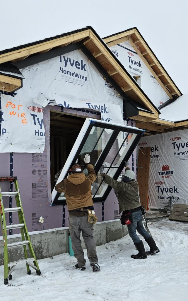 Two construction workers from Tobey Developments install a large window into the frame of a snow-covered house wrapped in Tyvek sheeting, with a green ladder propped nearby.