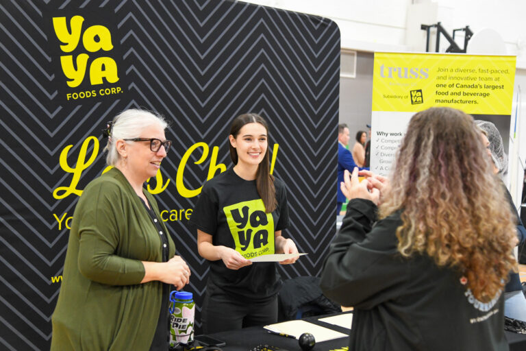 Two women stand at a Ya Ya Foods Belleville booth, smiling and talking to a visitor at a job fair. The booth features branded signs and informational materials about the company.