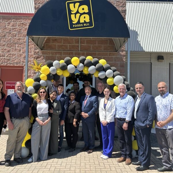 A group of people, including Bay of Quinte entrepreneurs, pose in front of a building entrance decorated with black, yellow, and white balloons under a “YaYa Foods B.V.” sign. The group is smiling, and it’s a sunny day.