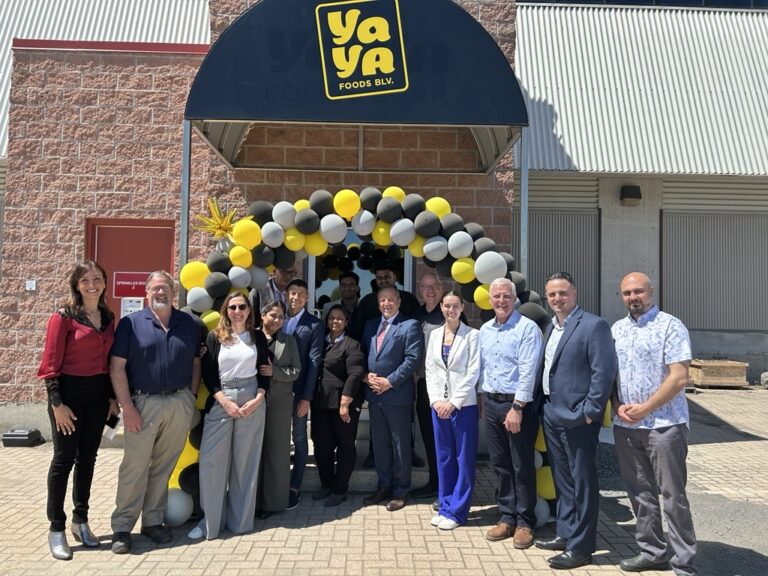 A group of people, including Bay of Quinte entrepreneurs, pose in front of a building entrance decorated with black, yellow, and white balloons under a “YaYa Foods B.V.” sign. The group is smiling, and it’s a sunny day.