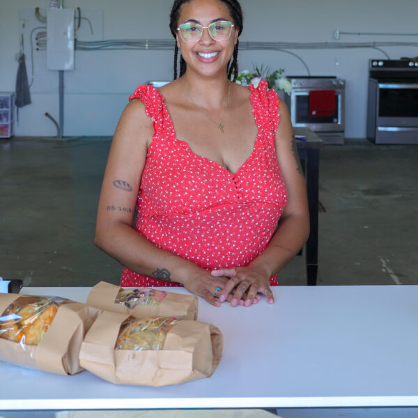 A smiling woman in a red dress stands behind a counter with three brown paper-wrapped sandwiches in a spacious, modern kitchen—capturing the spirit of Bay of Quinte entrepreneurs who infuse passion and creativity into every detail.