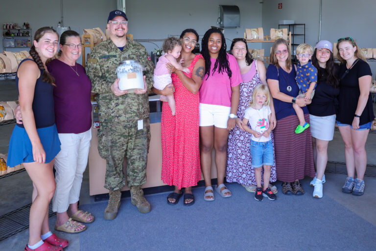 A group of eleven adults and three children smile and pose together indoors, with one adult in military uniform holding a jar. Among them are Bay of Quinte entrepreneurs, gathered proudly in front of shelves stocked with goods in the background.