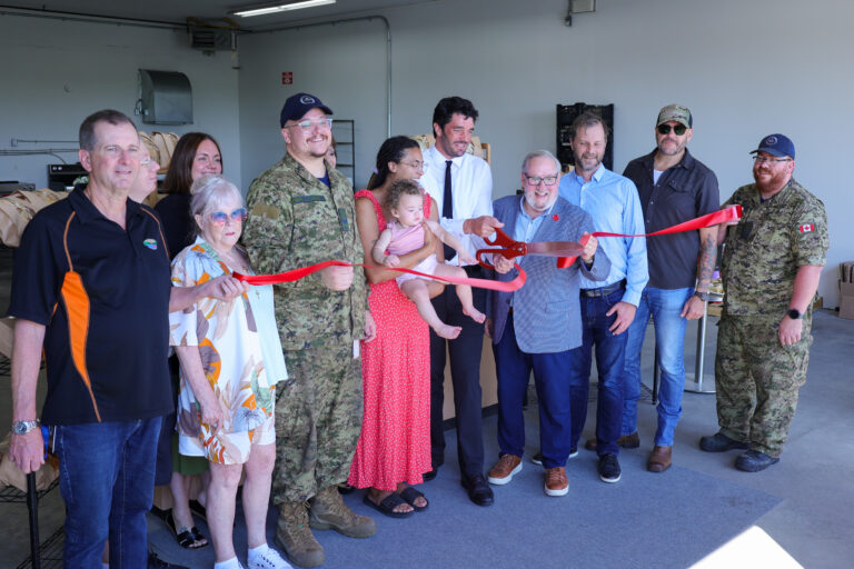 A group of people, including military personnel, civilians, and Bay of Quinte entrepreneurs, smile and cut a red ribbon at an indoor ribbon-cutting ceremony. One woman holds a child; everyone is dressed in casual or business attire.