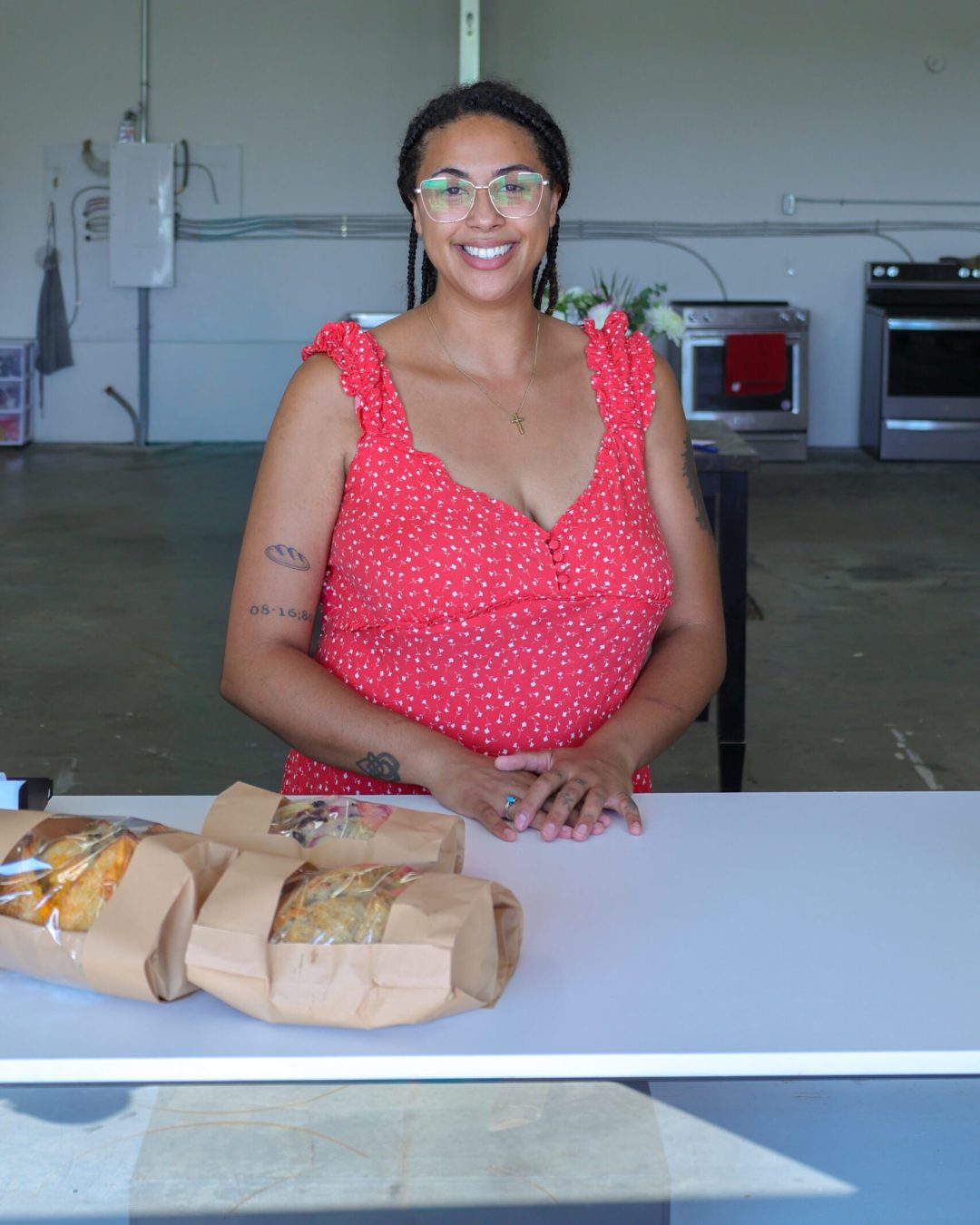 A smiling woman in a red dress stands behind a counter with three brown paper-wrapped sandwiches in a spacious, modern kitchen—capturing the spirit of Bay of Quinte entrepreneurs who infuse passion and creativity into every detail.